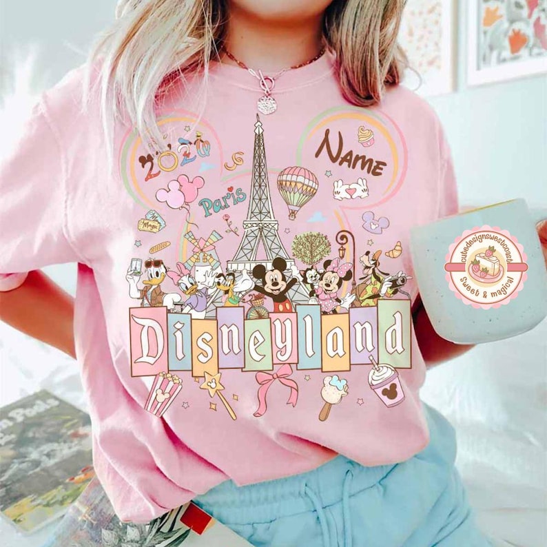 May include: Pink Disneyland t-shirt with a colorful graphic featuring Mickey and Minnie Mouse, Donald and Daisy Duck, and other Disney characters. The design includes the Eiffel Tower, a hot air balloon, and the word "Disneyland". A light blue mug with a dessert design is also visible.