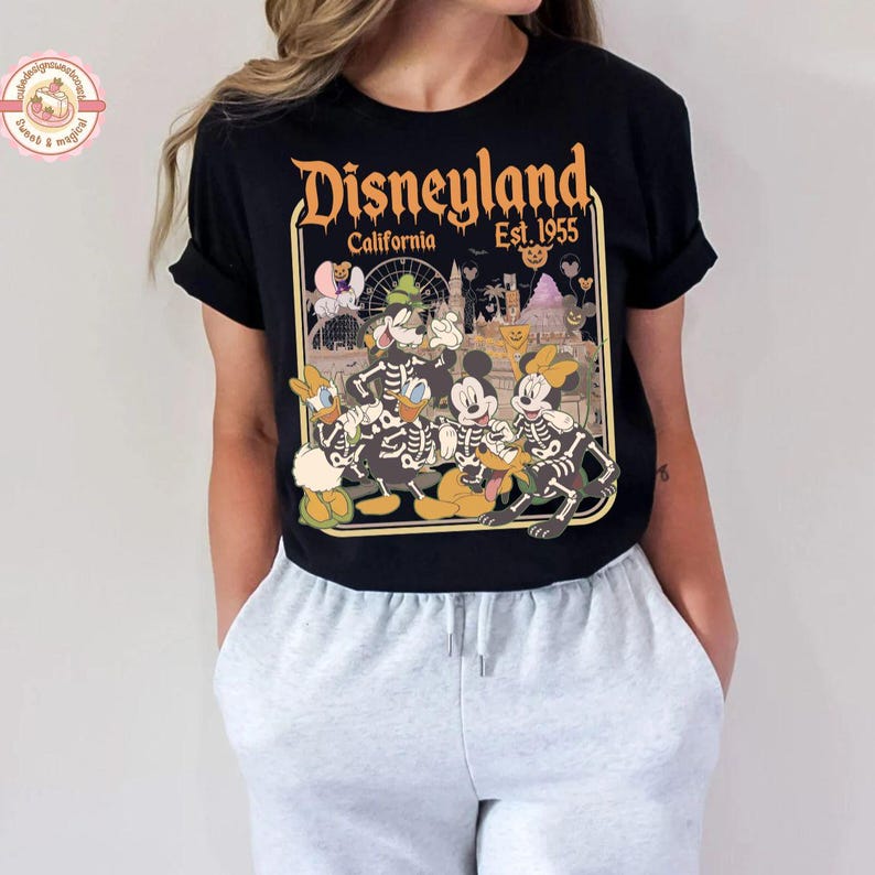 K&ouml;nnte beinhalten: Schwarzes T-Shirt mit einem Disneyland California-Motiv. Das Design zeigt Micky Maus, Donald Duck und andere Charaktere als Skelette. Der Text "Disneyland California Est. 1955" ist in Orange und Gelb gehalten.
