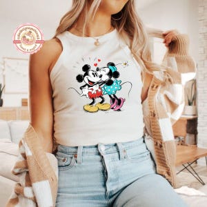 May include: A white tank top featuring a graphic of Mickey and Minnie Mouse embracing, with hearts and a flower. Mickey wears red shorts and yellow shoes, while Minnie wears a blue polka-dot dress and pink shoes. The top is worn by a person wearing blue jeans.