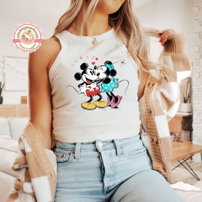 May include: A white tank top featuring a graphic of Mickey and Minnie Mouse embracing, with hearts and a flower. Mickey wears red shorts and yellow shoes, while Minnie wears a blue polka-dot dress and pink shoes. The top is worn by a person wearing blue jeans.