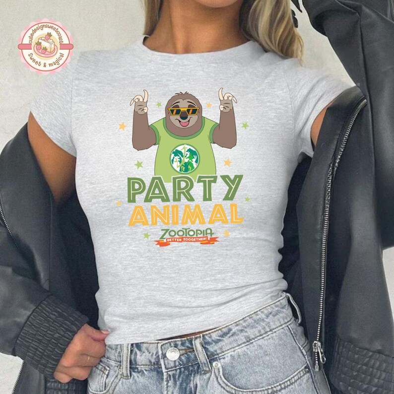May include: A light grey t-shirt with a cartoon sloth wearing sunglasses and a green shirt. The text "PARTY ANIMAL" is printed in green and yellow, with "ZOOTOPIA" below. The shirt has a crew neck and short sleeves.