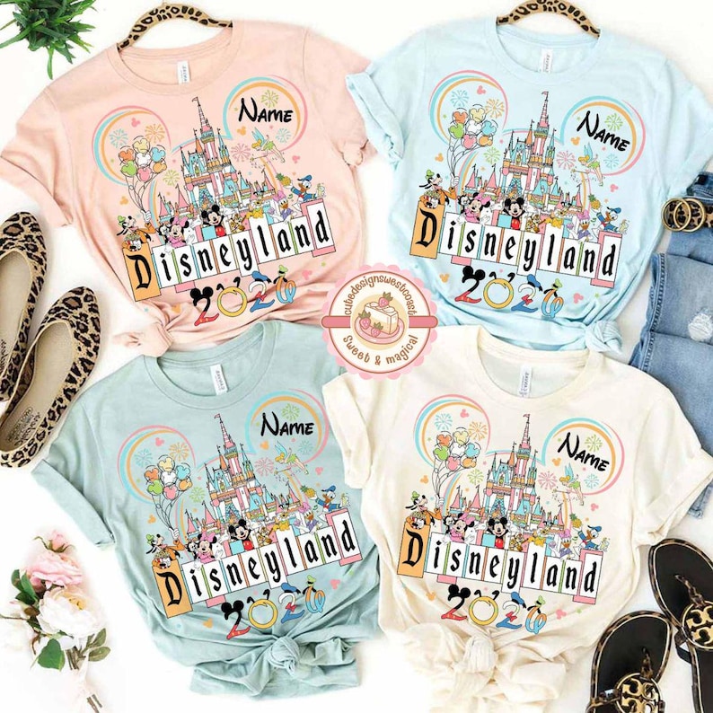 May include: Four pastel-colored t-shirts with a Disneyland graphic. Each shirt features a castle, balloons, and Disney characters. The word "Disneyland" and the year "2020" are printed on the front. The shirts are light pink, light blue, mint green, and cream.