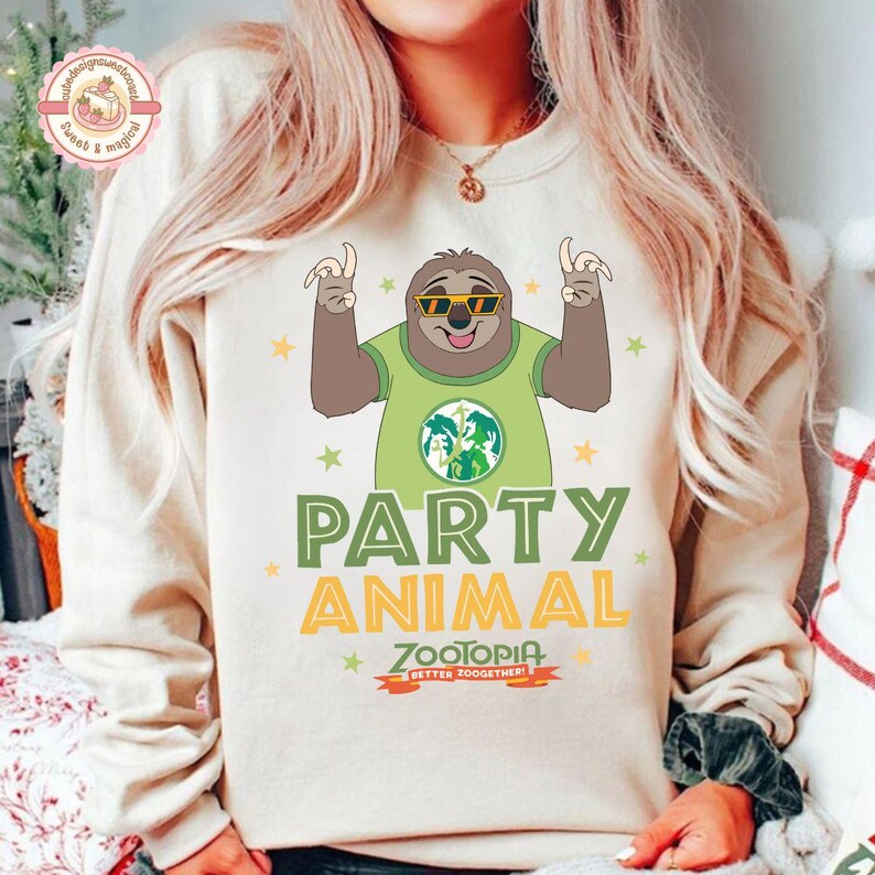 May include: Cream-coloured sweatshirt with a cartoon sloth wearing sunglasses and a green shirt with a globe design. The words "PARTY ANIMAL" and "ZOOTOPIA" are printed below the sloth, with the phrase "BETTER ZOOOGETHER!" at the bottom.