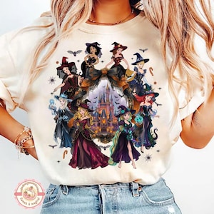 May include: A cream-colored t-shirt featuring a graphic print of various animated princesses dressed in Halloween-themed witch costumes. The design includes a central castle illustration framed by a large bow, surrounded by bats and spiderwebs.