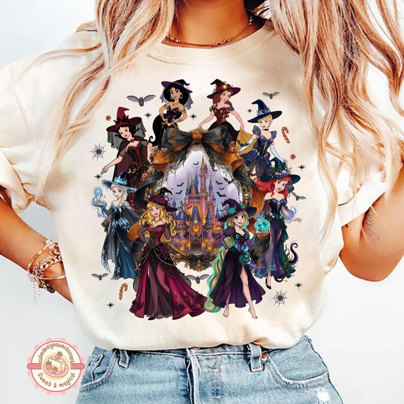 May include: A cream-colored t-shirt featuring a graphic print of various animated princesses dressed in Halloween-themed witch costumes. The design includes a central castle illustration framed by a large bow, surrounded by bats and spiderwebs.
