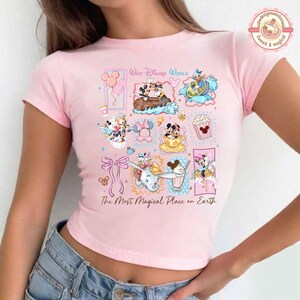 May include: A light pink t-shirt featuring a colorful graphic of various Disney characters and icons. The design includes Mickey and Minnie Mouse, Donald Duck, and Dumbo, along with the text "Walt Disney World" and "The Most Magical Place on Earth."