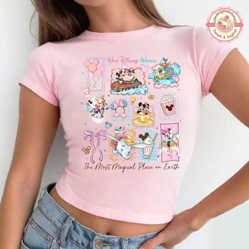 May include: A light pink t-shirt featuring a colorful graphic of various Disney characters and icons. The design includes Mickey and Minnie Mouse, Donald Duck, and Dumbo, along with the text "Walt Disney World" and "The Most Magical Place on Earth."