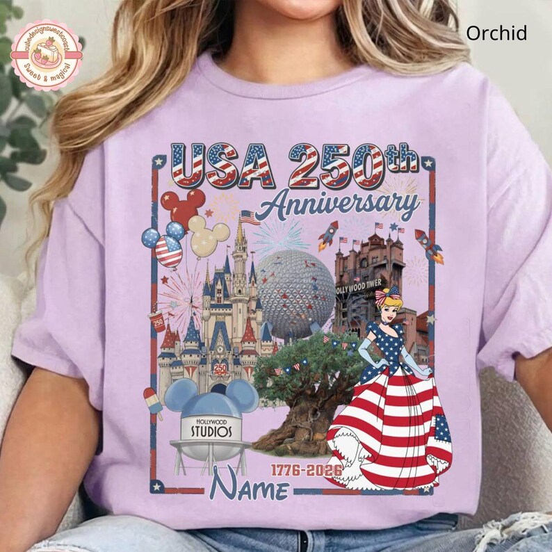 May include: Orchid-colored t-shirt with a graphic celebrating the USA's 250th Anniversary. The design includes iconic landmarks, fireworks, and a patriotic Cinderella in a red, white, and blue dress. The text "USA 250th Anniversary" is prominently displayed.