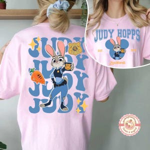May include: A light pink t-shirt featuring a cartoon image of Judy Hopps from Zootopia. The design includes the text "JUDY HOPPS" in blue, a police badge, and a carrot. The shirt also has the text "ZOOTOPIA".