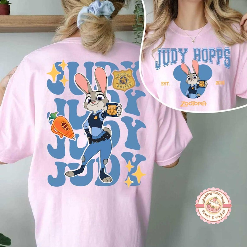 May include: A light pink t-shirt featuring a cartoon image of Judy Hopps from Zootopia. The design includes the text "JUDY HOPPS" in blue, a police badge, and a carrot. The shirt also has the text "ZOOTOPIA".