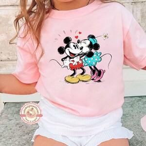 May include: A light pink t-shirt featuring a graphic of Mickey and Minnie Mouse kissing, with hearts above them. Mickey is wearing red shorts and yellow shoes, while Minnie wears a blue polka-dot dress and pink shoes. The shirt has short sleeves.