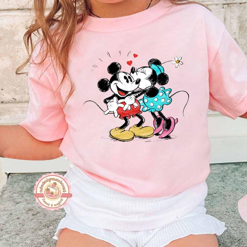 May include: A light pink t-shirt featuring a graphic of Mickey and Minnie Mouse kissing, with hearts above them. Mickey is wearing red shorts and yellow shoes, while Minnie wears a blue polka-dot dress and pink shoes. The shirt has short sleeves.