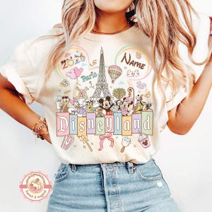 May include: Cream-colored t-shirt featuring a colorful Disneyland design. The design includes the Eiffel Tower, Mickey and Minnie Mouse, and other Disney characters. The word "Disneyland" is written in pastel colors.