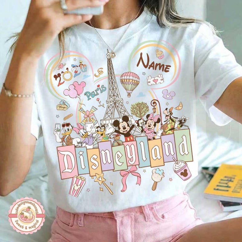 May include: White t-shirt featuring a colorful Disneyland design with Mickey and Minnie Mouse, Donald Duck, and other characters. The shirt includes the text "Disneyland" and "Paris" with a 2020 graphic.