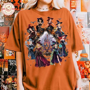 May include: An orange short-sleeve t-shirt featuring a graphic print of multiple animated characters dressed in witch costumes surrounding a central castle illustration, accented with bats, spiderwebs, and a large dark bow at the top.