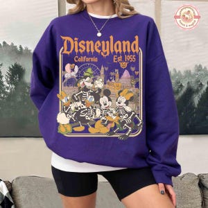 K&ouml;nnte beinhalten: Ein lila Disneyland-Sweatshirt mit einem Halloween-Motiv. Die Grafik zeigt Micky Maus, Minnie Maus, Donald Duck und Goofy als Skelette, mit dem Disneyland-Logo und dem Text "California Est. 1955".