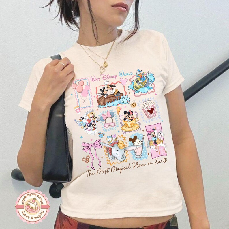 May include: Cream-colored t-shirt featuring colorful Disney World graphics. The shirt displays various Disney characters and icons, including Mickey and Minnie Mouse, Donald Duck, and Dumbo. Text on the shirt reads "Walt Disney World" and "The Most Magical Place on Earth."