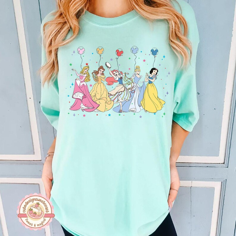 May include: A light blue t-shirt with a graphic of six Disney princesses holding balloons. The princesses are wearing their signature dresses and are surrounded by stars.