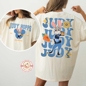 May include: Cream-colored t-shirt featuring a cartoon image of Judy Hopps from Zootopia. The front design includes the text "JUDY HOPPS" and a cartoon image of Judy. The back of the shirt has a larger image of Judy Hopps with the text "JUDY".