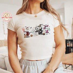 May include: A cream-coloured, short-sleeved t-shirt with a colourful graphic of characters from The Nightmare Before Christmas. The design includes Jack Skellington, Sally, and other Halloween Town residents. The shirt has a round neckline.