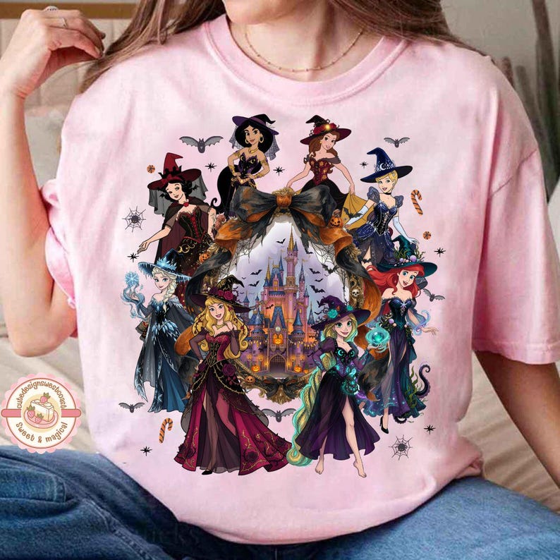 May include: A light pink t-shirt featuring a graphic print of various animated princesses dressed in witch costumes. The design includes a central castle illustration framed by a large bow, surrounded by bats, spiderwebs, and candy canes.