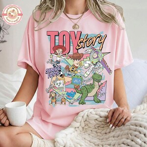 Pu&ograve; includere: Una t-shirt rosa chiaro con una stampa grafica colorata dei personaggi di Toy Story, tra cui Woody, Buzz Lightyear, Rex, Slinky Dog e Jessie. Il testo recita Toy Story Since 1995, Smarty Pants, Woody, Hi! Lilypad e Rex. La t-shirt ha maniche corte.