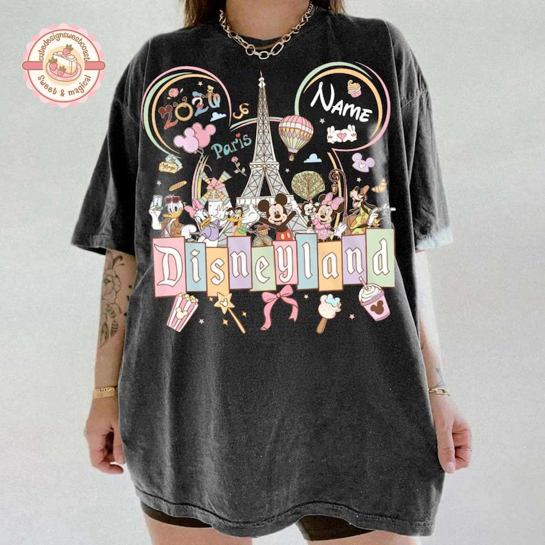 May include: Dark gray t-shirt featuring a colorful Disneyland Paris design. The graphic includes the Eiffel Tower, Mickey Mouse, and other Disney characters. The word "Disneyland" is prominently displayed.