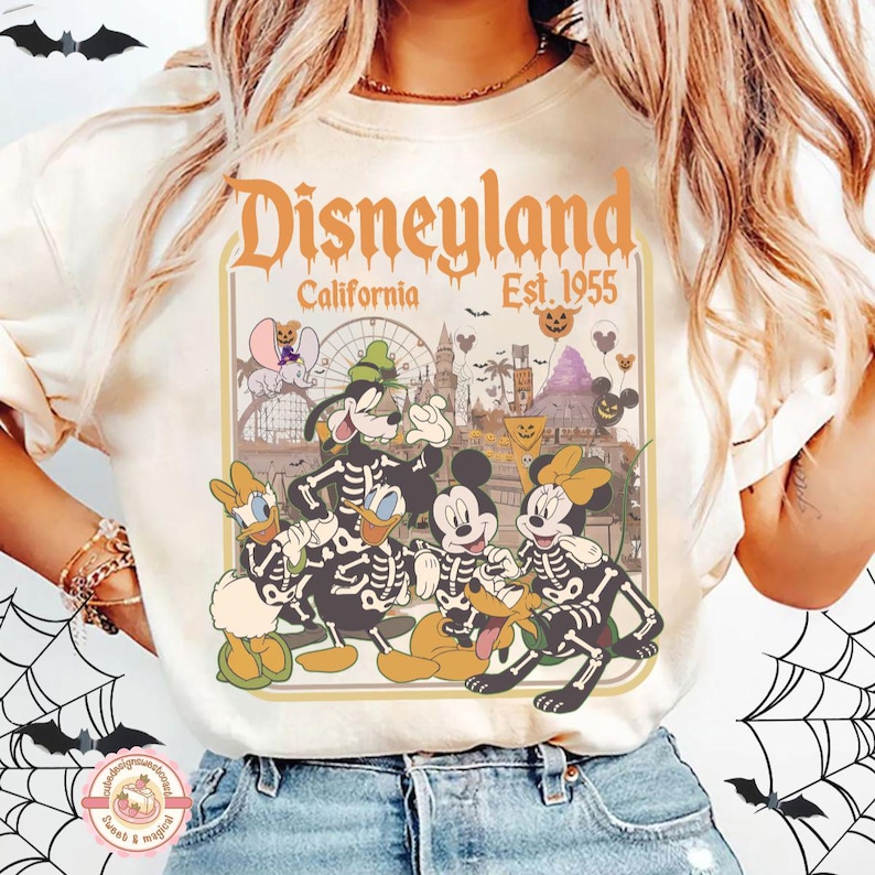 K&ouml;nnte beinhalten: Cremefarbenes T-Shirt mit Disneyland-Design und dem Text "Disneyland California Est. 1955". Die Grafik zeigt Disney-Figuren in Skelett-Kost&uuml;men mit Halloween-Thema.