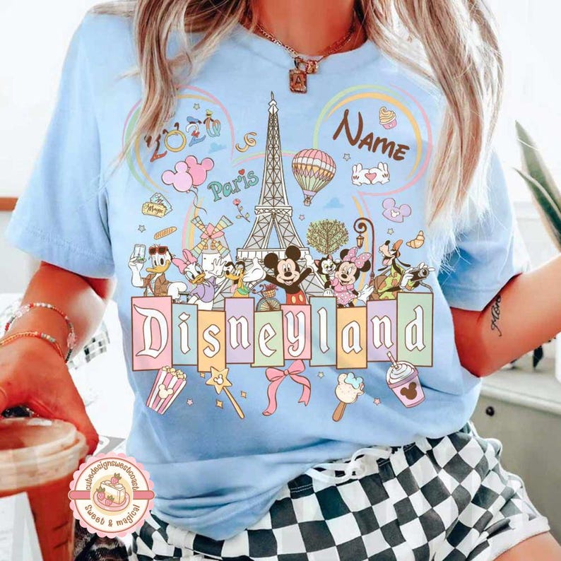 May include: Light blue t-shirt featuring the word "Disneyland" in pastel colors, with cartoon characters and the Eiffel Tower. The shirt has a design with the word "Paris" and the word "Name".