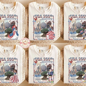 May include: Off-white t-shirts with a colorful graphic celebrating the USA's 250th Anniversary. Each shirt features a different cartoon character, the text "USA 250th Anniversary", and the years 1776-2026.
