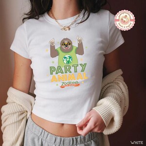 May include: White t-shirt with a cartoon sloth wearing sunglasses and a green shirt with a globe design. The shirt has the words "PARTY ANIMAL" and "Zootopia" printed on it. The t-shirt is a crew neck style.