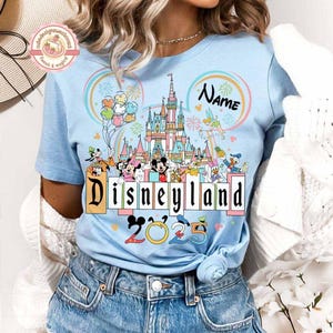 May include: A light blue t-shirt with a colorful illustration of the Disneyland castle and Mickey Mouse characters. The text "Disneyland 2023" is printed on the shirt. The shirt also has a pink circle with the word "Name" in it.