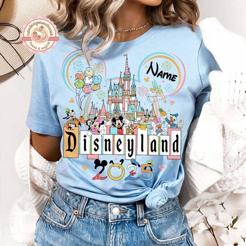 May include: A light blue t-shirt with a colorful illustration of the Disneyland castle and Mickey Mouse characters. The text "Disneyland 2023" is printed on the shirt. The shirt also has a pink circle with the word "Name" in it.