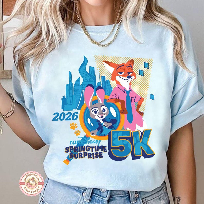 May include: Pale blue t-shirt with a graphic of Zootopia characters Judy Hopps and Nick Wilde, the text "2026" and "5K". The design includes the words "run Disney Springtime Surprise".