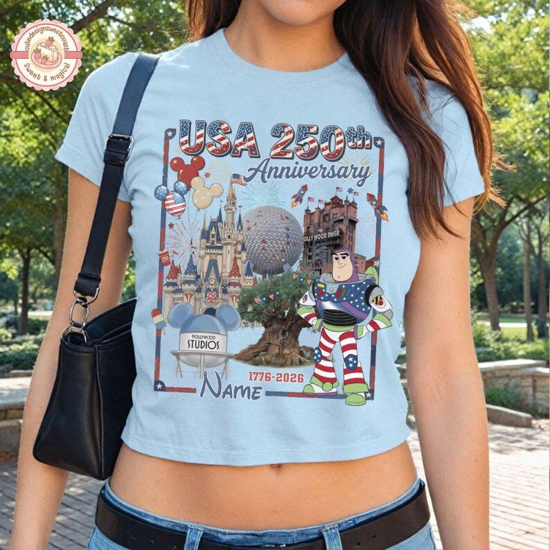 May include: Light blue t-shirt featuring a colorful graphic celebrating the USA's 250th Anniversary. The design includes iconic landmarks, balloons, rockets, and a Buzz Lightyear character. The text "USA 250th Anniversary" is prominently displayed.