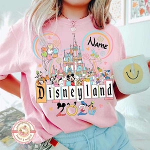 May include: Pink Disneyland 2020 t-shirt featuring a colorful graphic of the Disneyland castle, Mickey Mouse, Minnie Mouse, and other Disney characters. The shirt also has a rainbow design and the word "Name" above the castle.
