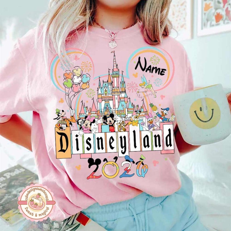 May include: Pink Disneyland 2020 t-shirt featuring a colorful graphic of the Disneyland castle, Mickey Mouse, Minnie Mouse, and other Disney characters. The shirt also has a rainbow design and the word "Name" above the castle.