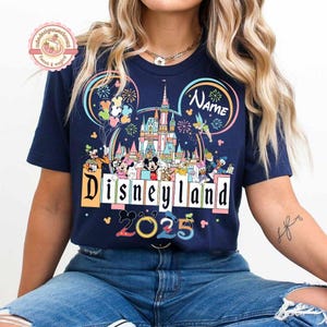 May include: A navy blue t-shirt with a colorful graphic of Mickey Mouse ears, fireworks, and the Disneyland castle. The text "Disneyland" and "2025" are printed on the shirt. The text "Name" is also printed on the shirt.