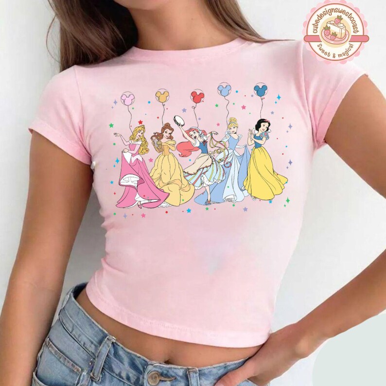 May include: Pink short-sleeved crop top with a graphic print of Disney princesses holding balloons. The princesses are wearing their signature dresses and have their hair styled in their iconic hairstyles. The text "cute designs sweet concepts sweet & magical" is printed in the top right corner of the image.
