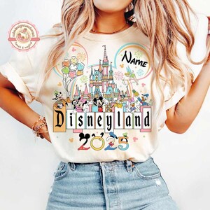 May include: A white t-shirt with a colorful graphic of Mickey Mouse and other Disney characters in front of Cinderella's Castle. The text "Disneyland 2023" is printed below the graphic. The text "Name" is printed above the castle.