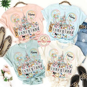 May include: Four pastel colored t-shirts with a graphic of Cinderella's castle and Mickey Mouse characters. The shirts say "Disneyland 2023" and have the word "Name" on them.