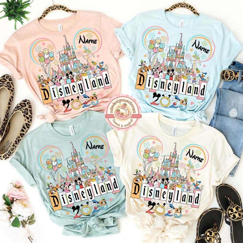 May include: Four pastel colored t-shirts with a graphic of Cinderella's castle and Mickey Mouse characters. The shirts say "Disneyland 2023" and have the word "Name" on them.