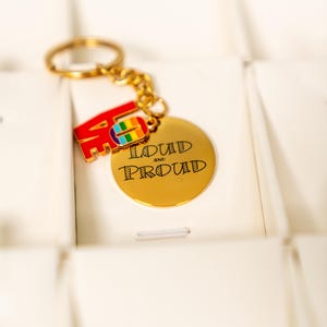 May include: A gold-colored keychain with a round charm that reads "LOUD AND PROUD" in black lettering. The keychain also features a red "LOVE" charm with a rainbow-colored hand symbol. The keychain is attached to a gold-colored keyring.