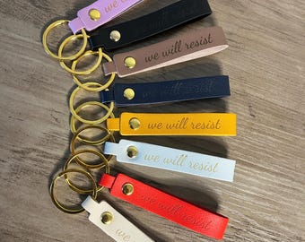 May include: Seven leather keychains with gold key rings. Each keychain has the text "we will resist" in gold lettering. The keychains are in different colors: pink, black, brown, navy blue, yellow, light blue, and white.