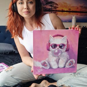 Może przedstawiać: Kobieta trzyma obraz przedstawiający białego kota w różowych słuchawkach i okularach przeciwsłonecznych, dmuchającego bańkę. Kot siedzi na różowym tle. Obraz jest oprawiony w ramkę z różową obwódką.