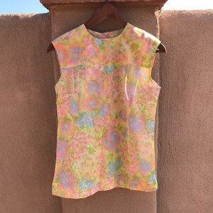 May include: A sleeveless top with a floral pattern in pastel colors. The top features a round neckline and a zipper closure. The fabric has a sheer overlay, and the floral design includes pink, blue, and green elements on a yellow background.