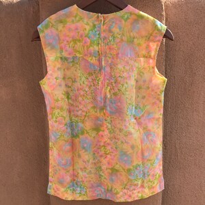 May include: A sleeveless floral top in pastel colors of pink, yellow, blue, and green. The vintage blouse has a zipper down the back and is hanging on a wooden hanger. The floral pattern is visible across the entire garment.