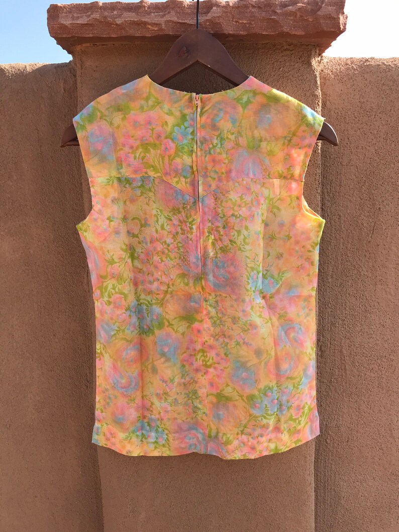 May include: A sleeveless floral top in pastel colors of pink, yellow, blue, and green. The vintage blouse has a zipper down the back and is hanging on a wooden hanger. The floral pattern is visible across the entire garment.