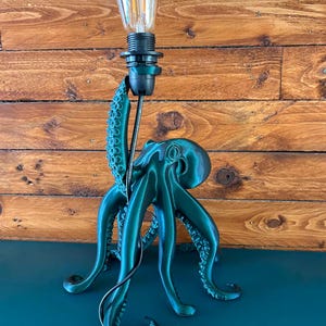 May include: A dark green octopus-shaped lamp base with a black socket and a clear glass Edison-style light bulb. The octopus has textured tentacles with visible suckers, and one tentacle extends upwards to support the light fixture and black cord.