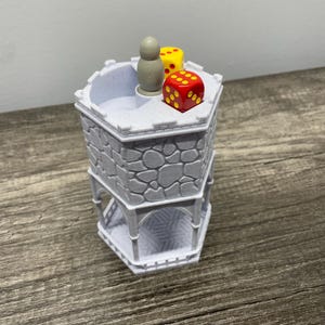 May include: A white tower-shaped dice rolling tower with a gray wooden figure and three dice on top. Two of the dice are red with yellow dots and one is yellow with red dots.